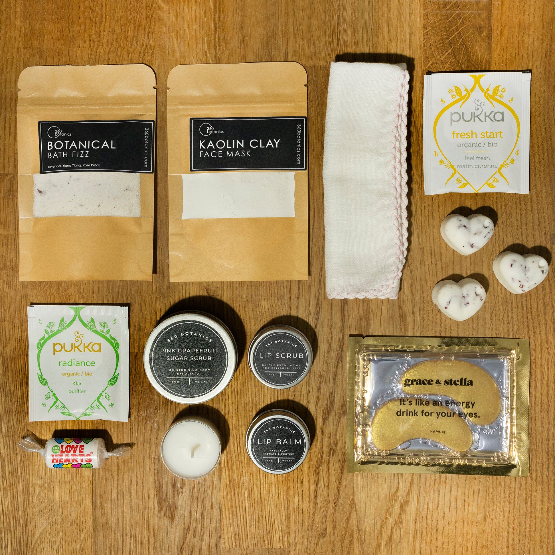 An array of self-care items neatly displayed on a wooden surface: two pouches labeled "Botanical Bath Fizz" and "Kaolin Clay Face Mask," a white washcloth, two Pukka tea packets in "fresh start" and "radiance" varieties, a tin of "Pink Grapefruit Sugar Scrub," two round containers labeled "Lip Scrub" and "Lip Balm," a small white candle, heart-shaped bath bombs, and a "Grace & Stella" eye mask packet.