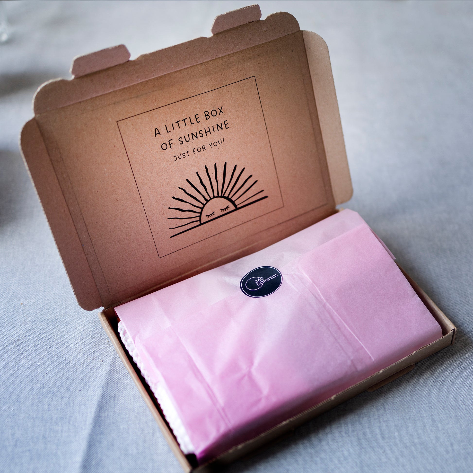 An open cardboard box with the phrase "A LITTLE BOX OF SUNSHINE JUST FOR YOU!" printed inside the lid, with a stylized sunburst design. The contents are wrapped in pink tissue paper, sealed with a round sticker bearing the 360 Botanics logo, set against a light gray fabric background