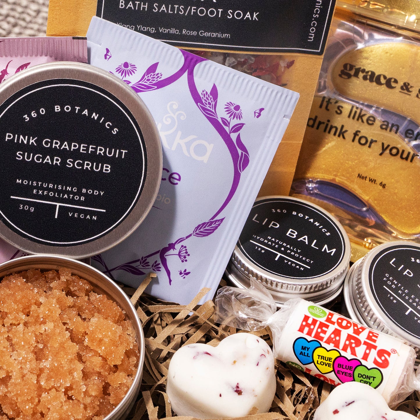 Close-up of a 360 Botanics gift set featuring a variety of skincare products. Visible items include Pink Grapefruit Sugar Scrub in an open tin revealing its texture, heart-shaped bath bombs, lip balm tins, and colorful Love Hearts candy. Packets of tea and bath salts with descriptions add to the indulgent self-care theme of the basket