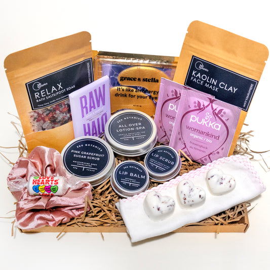 A curated 360 Botanics gift basket filled with a variety of wellness products. It includes bath salts, a kaolin clay face mask, organic tea, a raw chocolate bar, sugar scrub, all-over lotion, and lip balm, all nestled in natural straw. A pink satin hair scrunchie and heart-shaped bath bombs add a touch of femininity to the collection