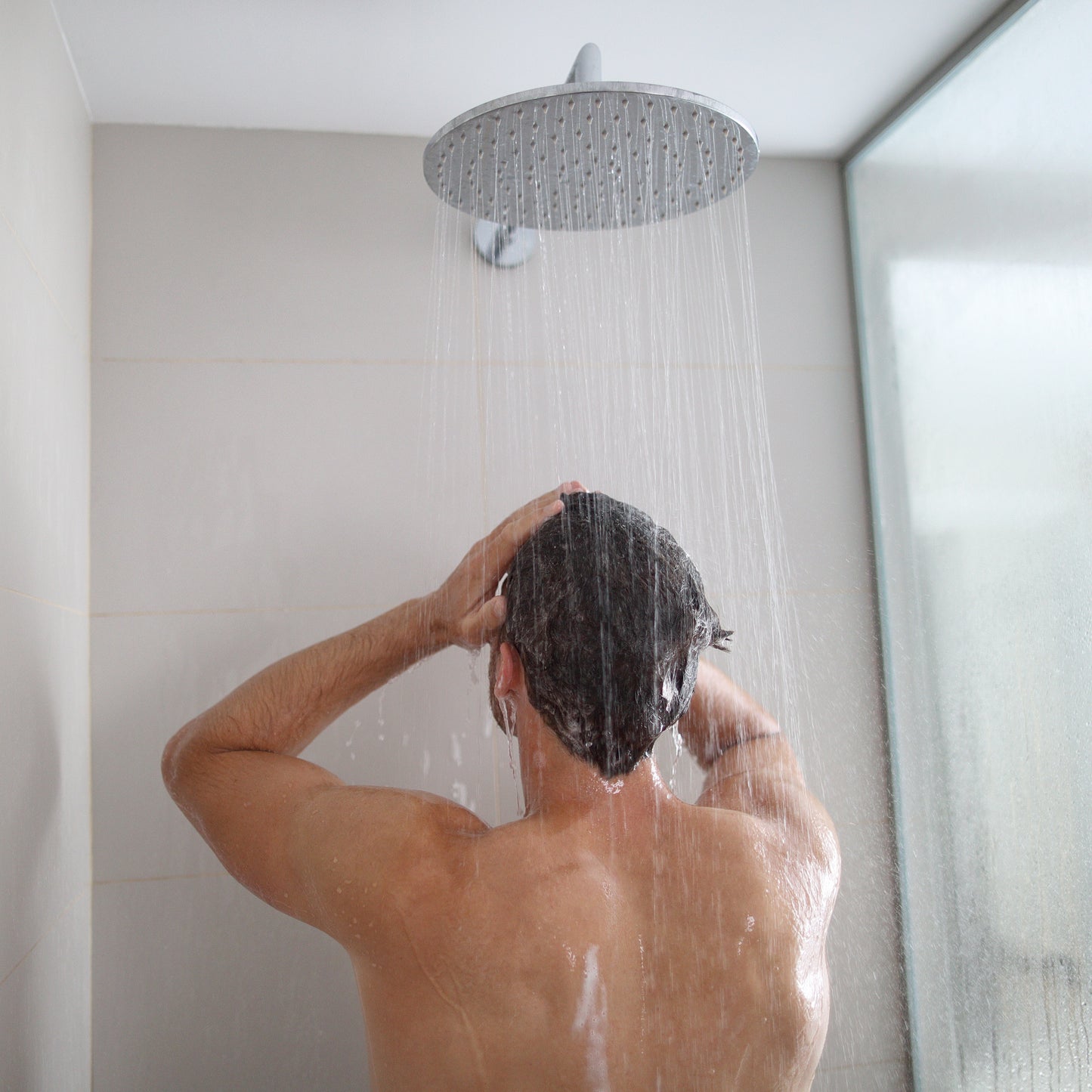 A man standing under a rainfall shower head in a modern bathroom, washing his hair with both hands as water streams down, creating a refreshing and serene moment
