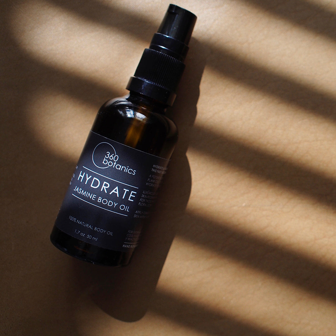 A bottle of 360 Botanics HYDRATE Jasmine Body Oil with a spray nozzle, placed diagonally across a textured background with dynamic light and shadow stripes cast by a window blind.