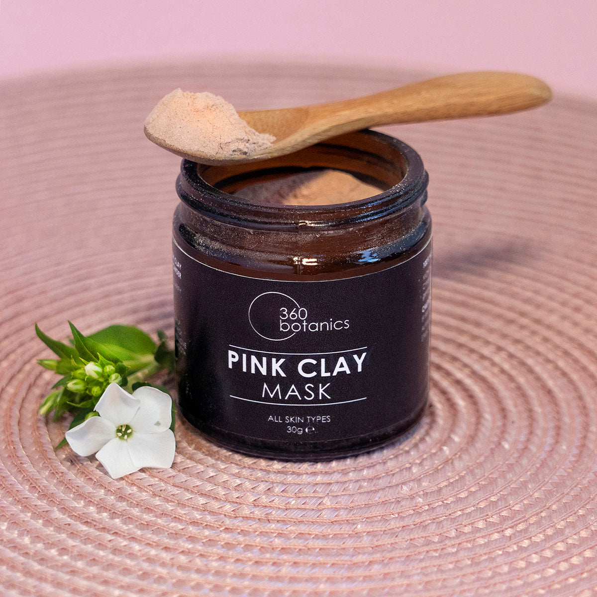 An open jar of 360 Botanics Pink Clay Mask is presented on a textured woven mat. A wooden spoon with a scoop of the fine pink clay powder rests on top, while a delicate white flower with green leaves adds a natural touch to the scene, emphasizing the organic beauty product suitable for all skin types