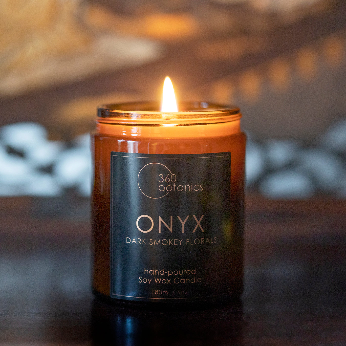 The 360 Botanics ONYX soy wax candle with 'Dark Smokey Florals' scent is featured, gently illuminating a room with its tranquil flame. The amber glass jar adds warmth to the setting, creating an inviting and serene ambiance, perfect for relaxation or as a sophisticated home decor element