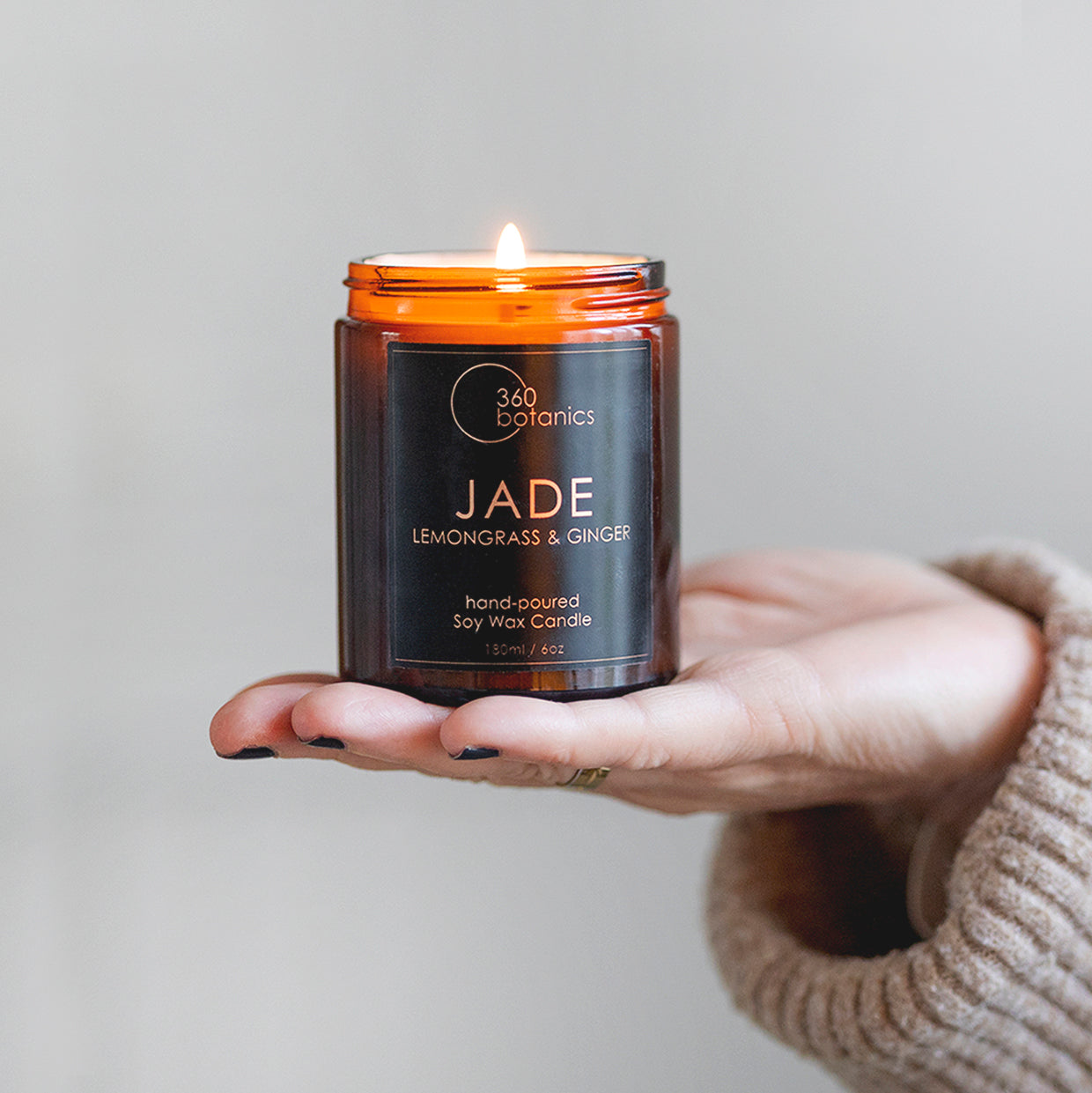 Jade candle in amber jar glowing held out on woman's handA person holds a lit soy wax candle by "360 botanics" named JADE with lemongrass and ginger scent. The candle is in a small amber glass jar with a label detailing the brand and scent, and the warm glow of the flame is visible at the top. The person is wearing a beige sweater, and only their hand is in the frame against a neutral background, highlighting the candle.
