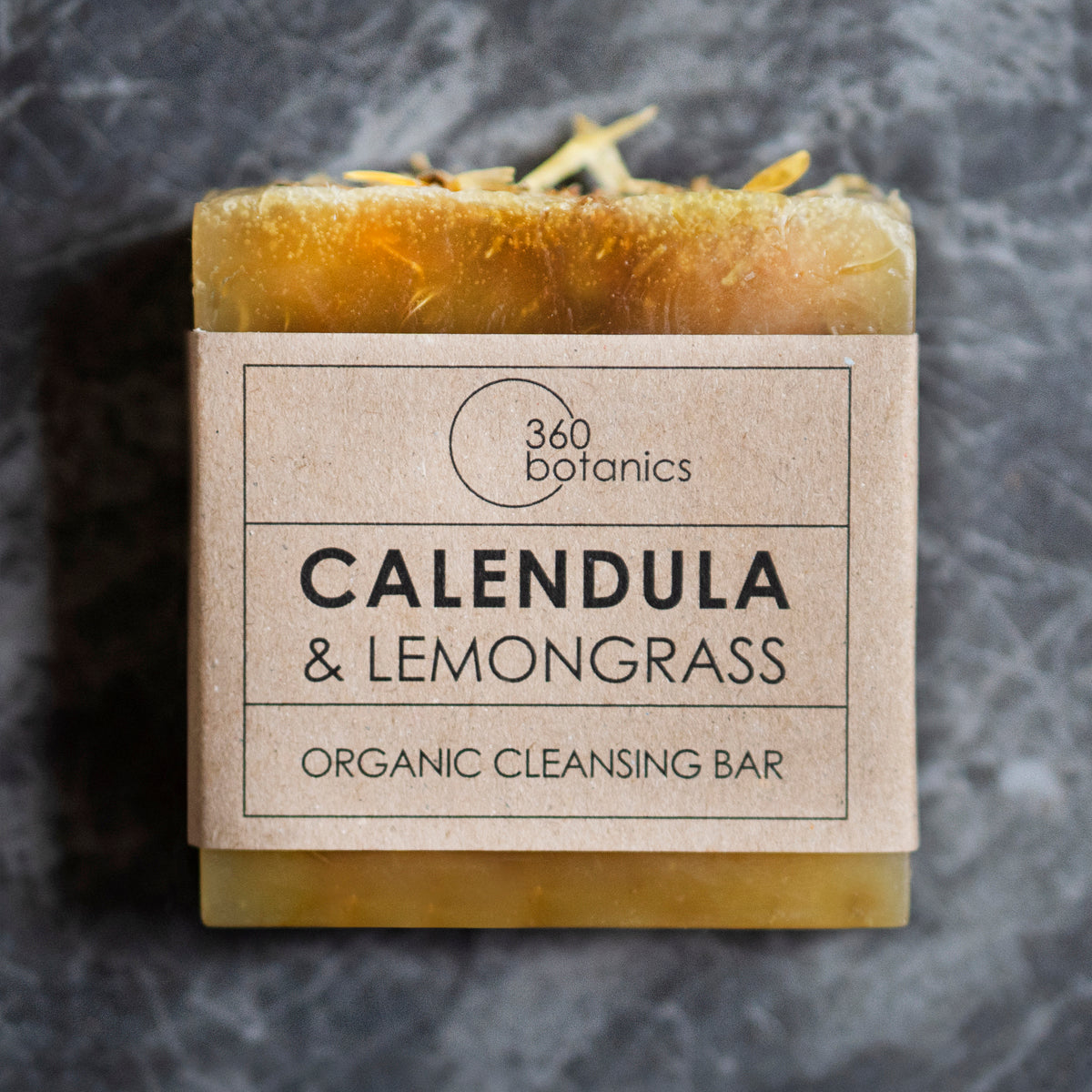 A bar of 360 Botanics organic cleansing soap labeled "CALENDULA & LEMONGRASS," wrapped in brown paper packaging and set against a marbled grey background.