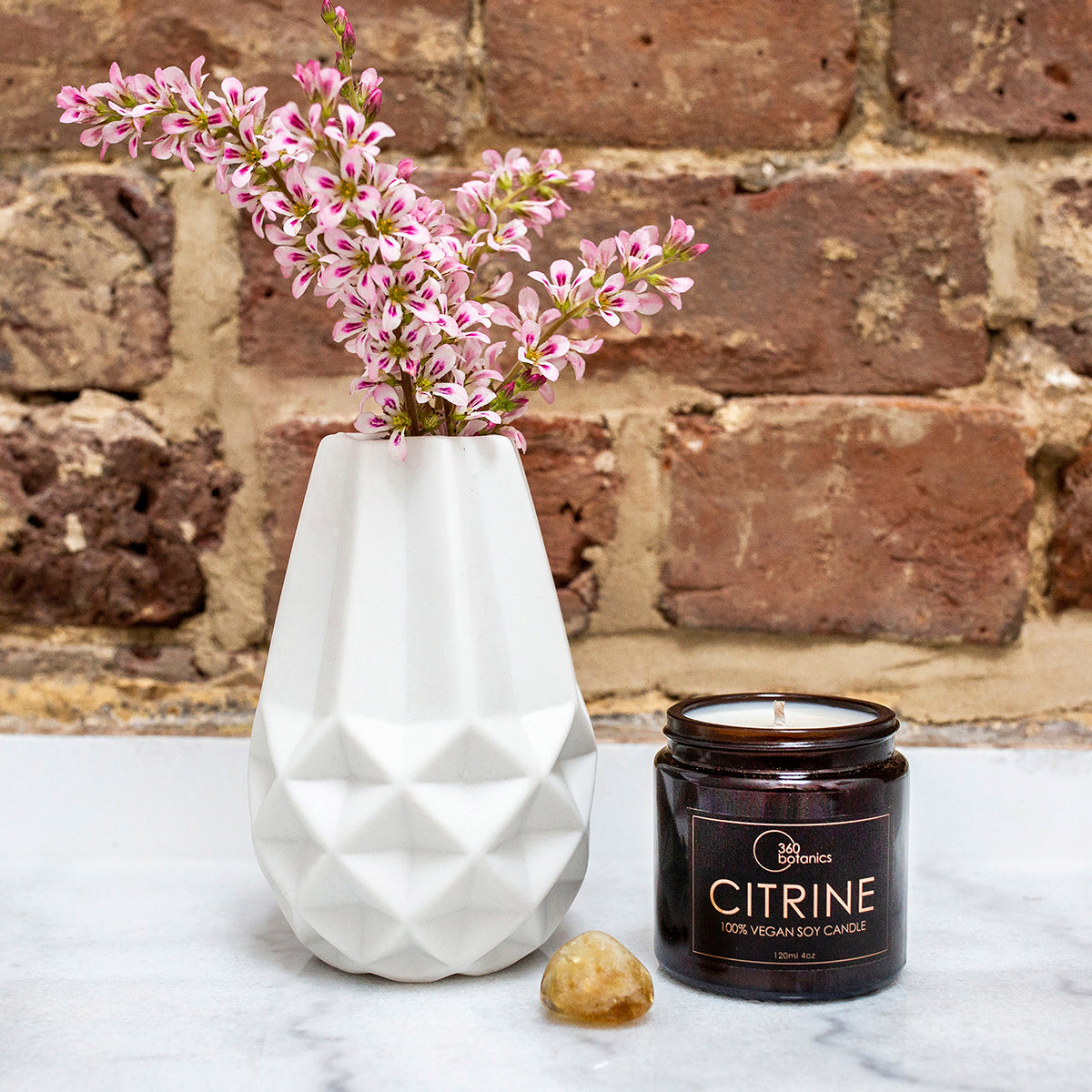 A white geometric vase with pink flowers next to an unlit 360 Botanics Citrine 100% vegan soy candle in a dark glass jar, accompanied by a citrine crystal, set against a brick wall on a white surface.