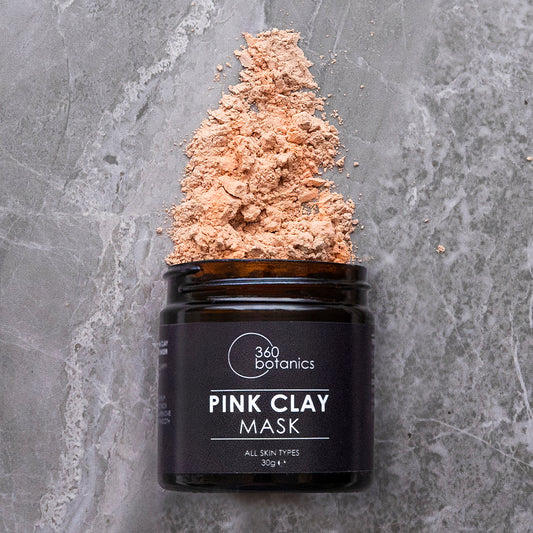 A jar of 360 Botanics Pink Clay Mask is open, with its powdered content artistically spilled on top to form a peak. The black jar contrasts against a gray marbled background, emphasizing the natural and pure ingredients suitable for all skin types as indicated on the label