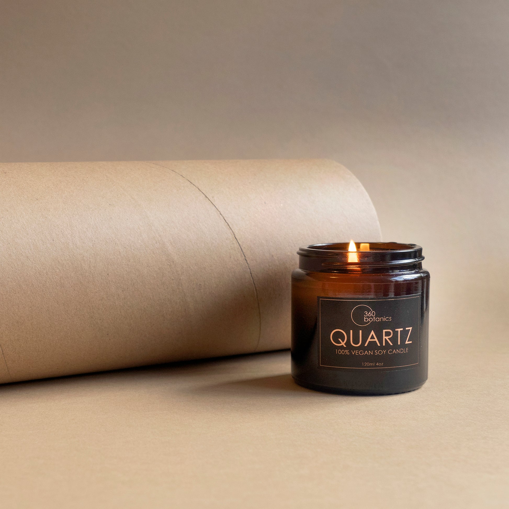  A lit soy candle in a brown glass jar is displayed against a craft paper backdrop that rolls off to one side. The candle's label reads "360 Botanics QUARTZ 100% Vegan Soy Candle, 120ml 4oz," illuminated by the soft glow of the flame, adding warmth to the minimalist setting.