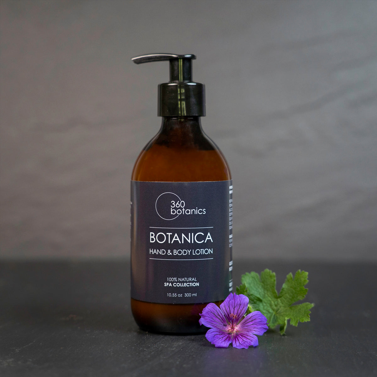 A bottle of 360 Botanics SPA All Over Lotion placed on a soft surface with a vibrant mauve Geranium flower next to it. The elegant design of the bottle reflects the lotion's luxurious and natural formulation.