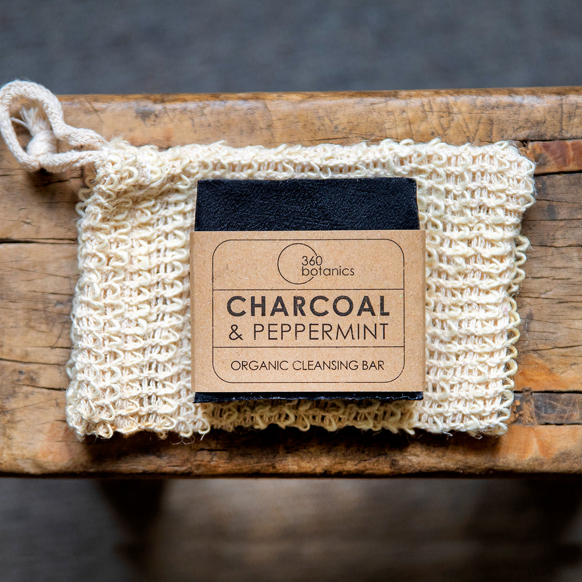 A 360 Botanics Charcoal & Peppermint Organic Cleansing Bar placed on a natural wooden surface, partially wrapped in a textured beige loofah with a hanging loop. The kraft paper label adds an eco-friendly touch, emphasizing the organic nature of the skincare product