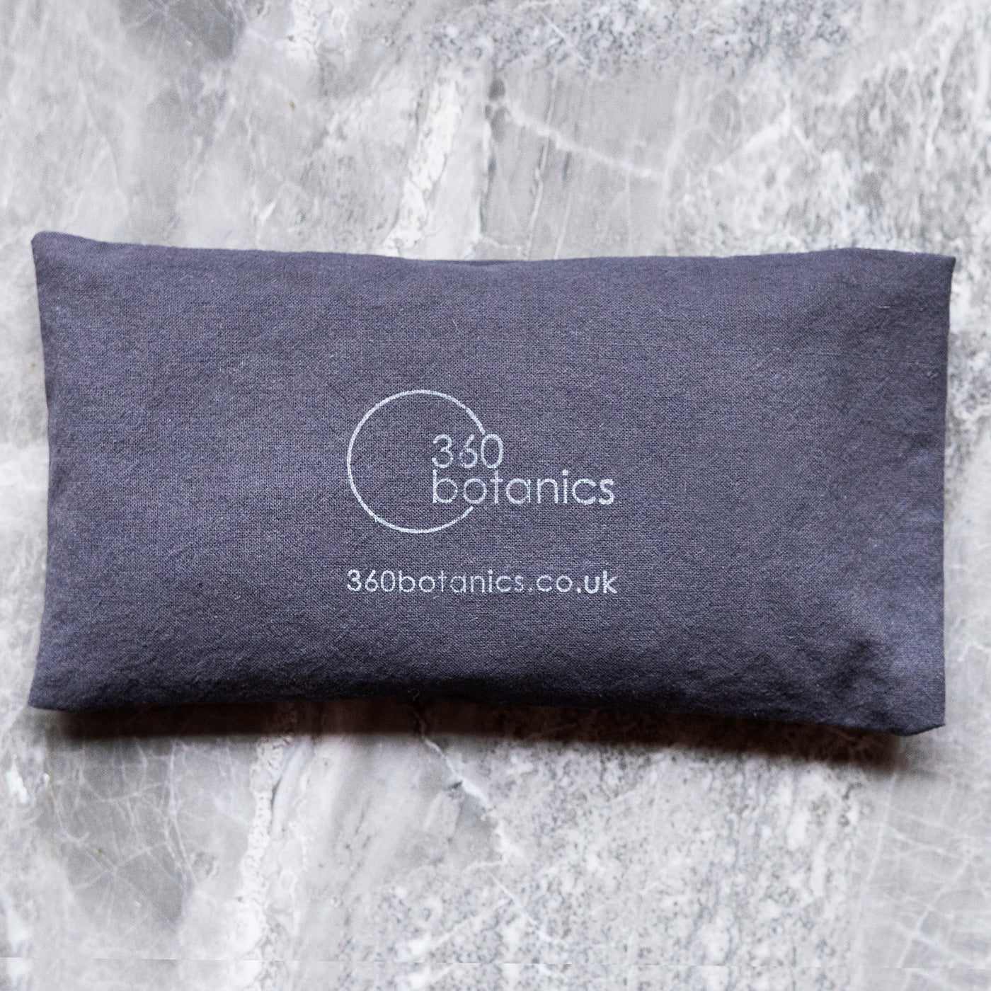 A navy blue eye pillow with the white logo "360 Botanics" and the website "360botanics.co.uk" on a marbled grey background.