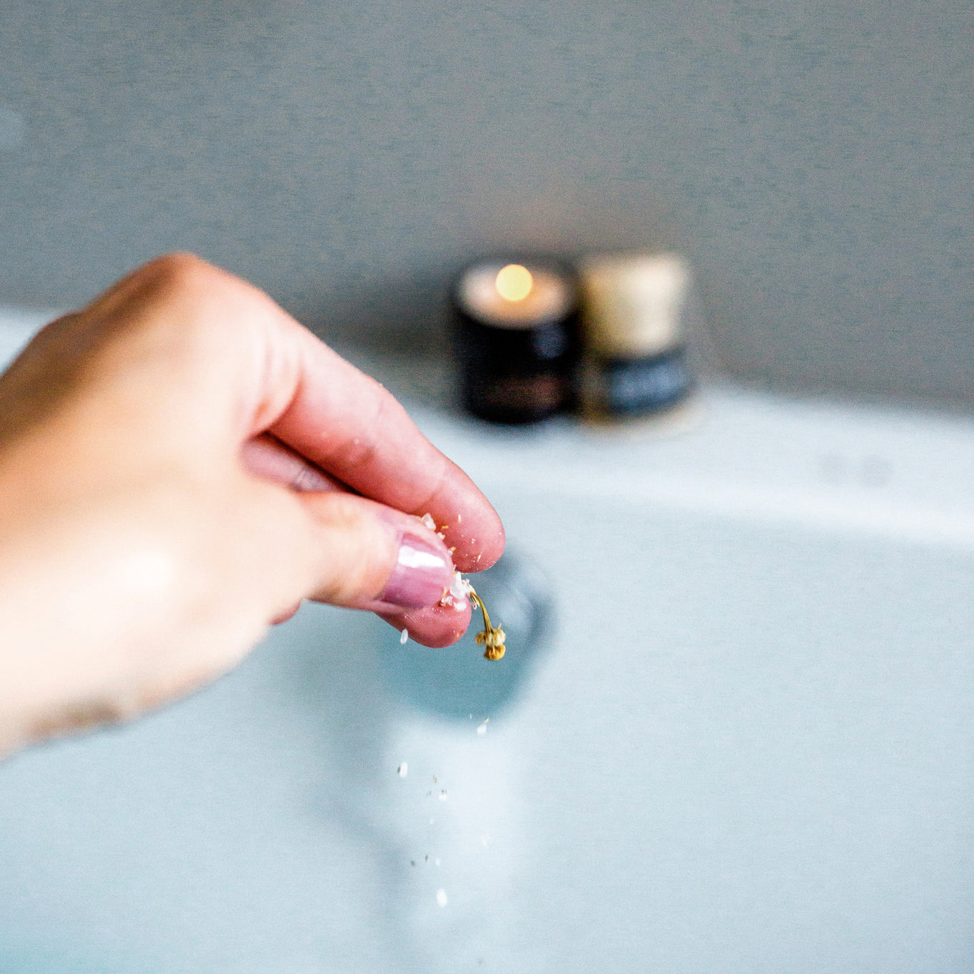 A hand with a light pink manicure sprinkles bath salts into a bathtub. The focus is on the fingers releasing small salt crystals, capturing the motion with a slight blur. In the soft-focused background, a candle burns, providing a serene and warm ambiance. The scene suggests a relaxing, spa-like atmosphere within a home bathroom.