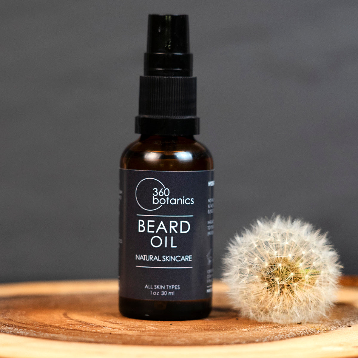 A bottle of 360 Botanics Beard Oil labeled for "All Skin Types" and "Natural Skincare" in a 1 oz 30 ml size with a spray nozzle, placed on a wooden surface next to a dandelion puff, against a blurred grey background