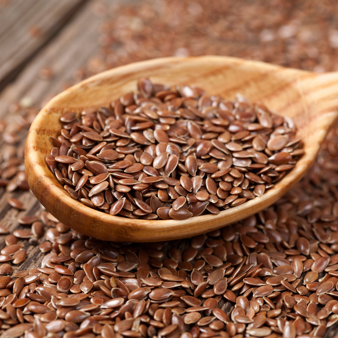 A wooden spoon overflowing with brown flaxseeds set on a backdrop of more flaxseeds on a rustic wooden surface.