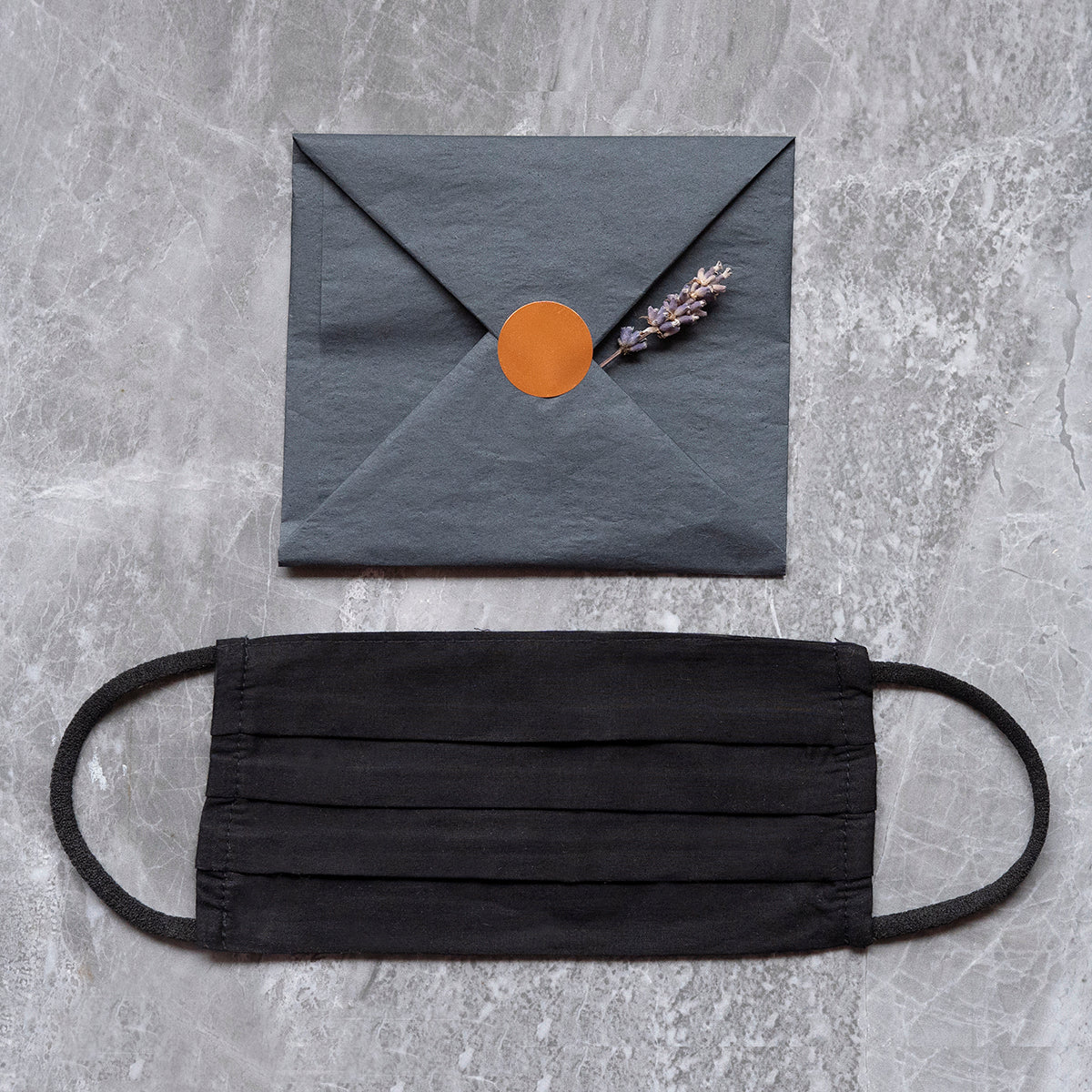 A black fabric face mask laid out on a marble surface below a dark grey envelope sealed with an orange sticker and adorned with a sprig of lavender.