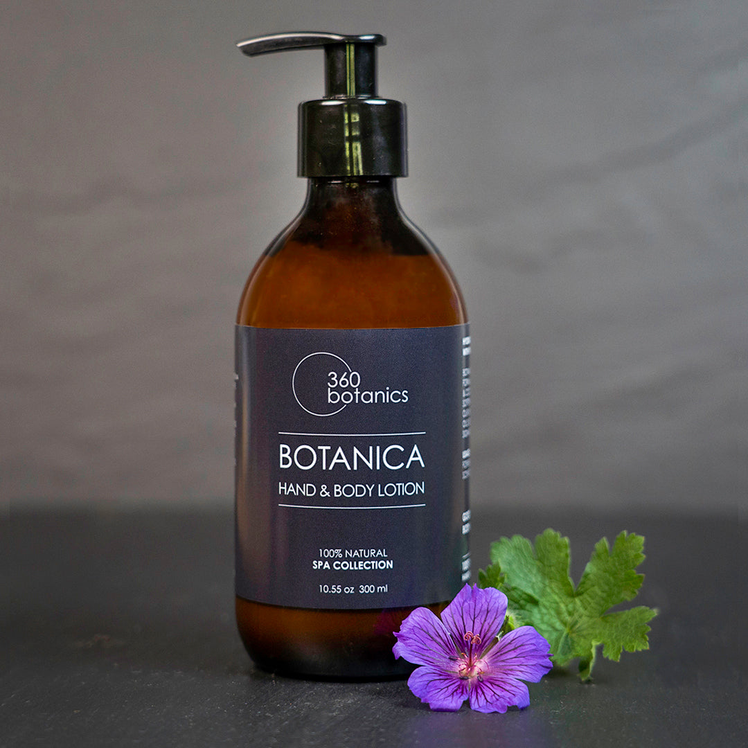 A bottle of 360 Botanics Botanica Hand & Body Lotion from the 100% Natural Spa Collection, sized at 10.55 oz or 300 ml, with a pump dispenser. Beside the bottle lies a purple flower and green leaves against a grey background.