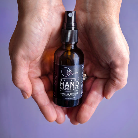 Cupped hands holding a bottle of 360 Botanics Citrus Hand Sanitiser with a spray nozzle, labeled as "Natural Defence Cleanse & Sanitise" in a 1.7 oz or 50 ml size, with a purple background