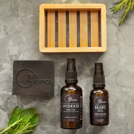 Two bottles of 360 Botanics skincare products on a gray marbled surface, one labeled 'HYDRATE BODY OIL' and the other 'BEARD OIL.' A black bar of soap with '360 Botanics' embossed on it lies to the left, and a bamboo tray rests in the background. Fresh green leaves are scattered around for a natural touch