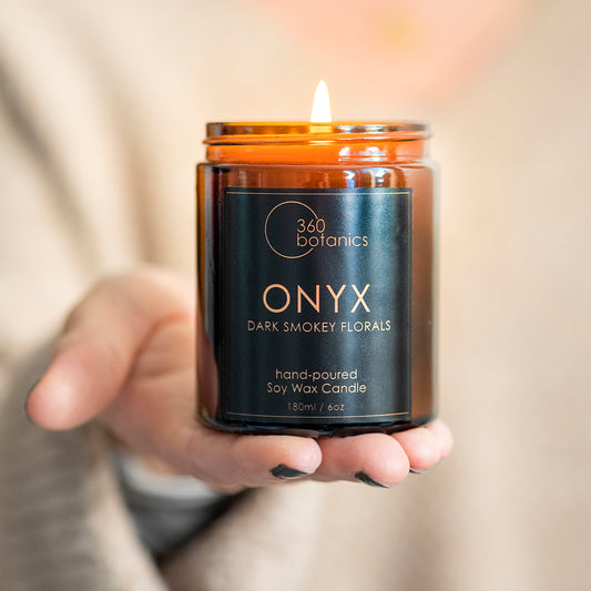 A hand holds a lit 360 Botanics ONYX soy wax candle with a label that reads 'Dark Smokey Florals'. The warm flame glows in an amber-colored glass container, conveying a cozy atmosphere and the candle's hand-poured quality, suitable for adding ambience and fragrance to any space