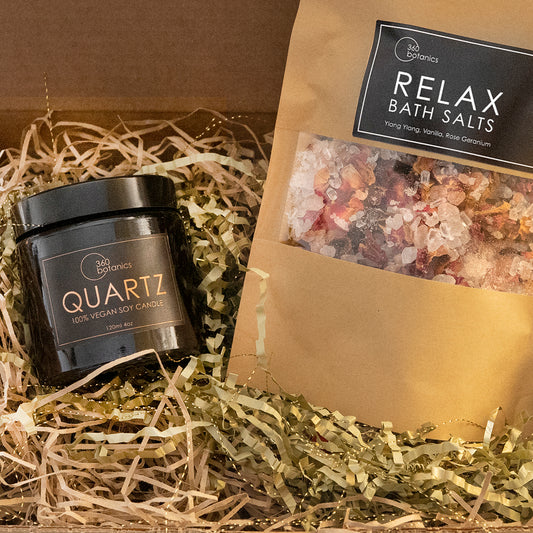 A cosy and inviting gift set from 360 Botanics, presented in a brown cardboard box filled with natural shredded paper. Inside, there's a sleek black glass jar labeled 'QUARTZ 100% VEGAN SOY CANDLE' and a kraft paper pouch of 'RELAX BATH SALTS' with visible crystals of Ylang Ylang, Vanilla, and Rose Geranium, promising a soothing and aromatic experience