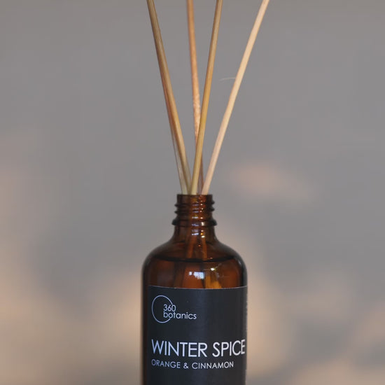 An atmospheric video of the 360 Botanics Winter Spice reed diffuser with orange and cinnamon, placed on a wooden coaster. The amber bottle with light brown reeds is highlighted by a soft, warm light in the background, invoking a cozy winter ambiance