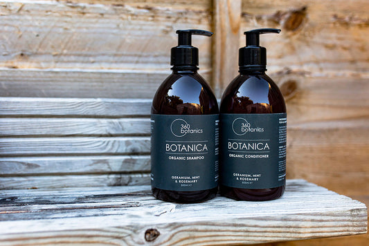 Two 500ml bottles of 360 Botanics Botanica products, one an organic shampoo and the other an organic conditioner, with geranium, mint, and rosemary scents, placed on a rustic wooden surface.