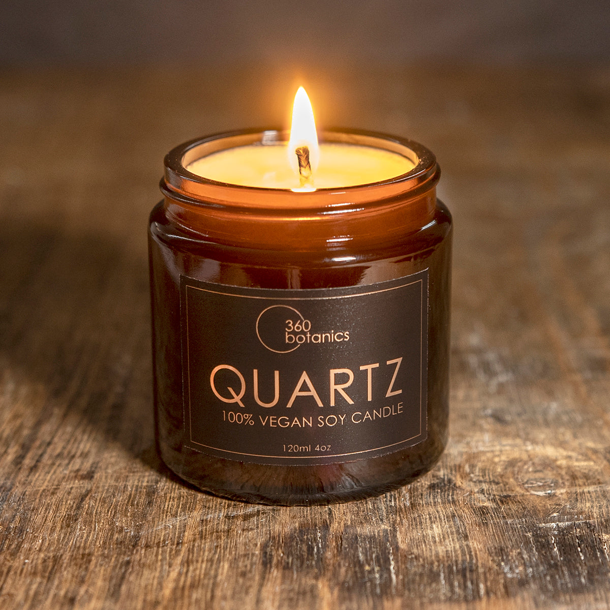 A lit soy candle with a single wick burns brightly within a small brown glass jar on a rustic wooden surface. The label on the jar clearly states "360 Botanics QUARTZ 100% Vegan Soy Candle, 120ml 4oz", highlighting the candle's brand and vegan quality. The warm light of the candle casts a soft glow on the wood, enhancing the cozy atmosphere.
