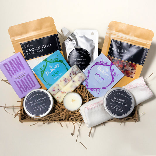 open cardboard gift box with two kraft pouches, two herbal tea bags, wax melts, a chocolate bar with lilac-coloured packaging , a t-light candle, silver sachet containing all over body wash, white background