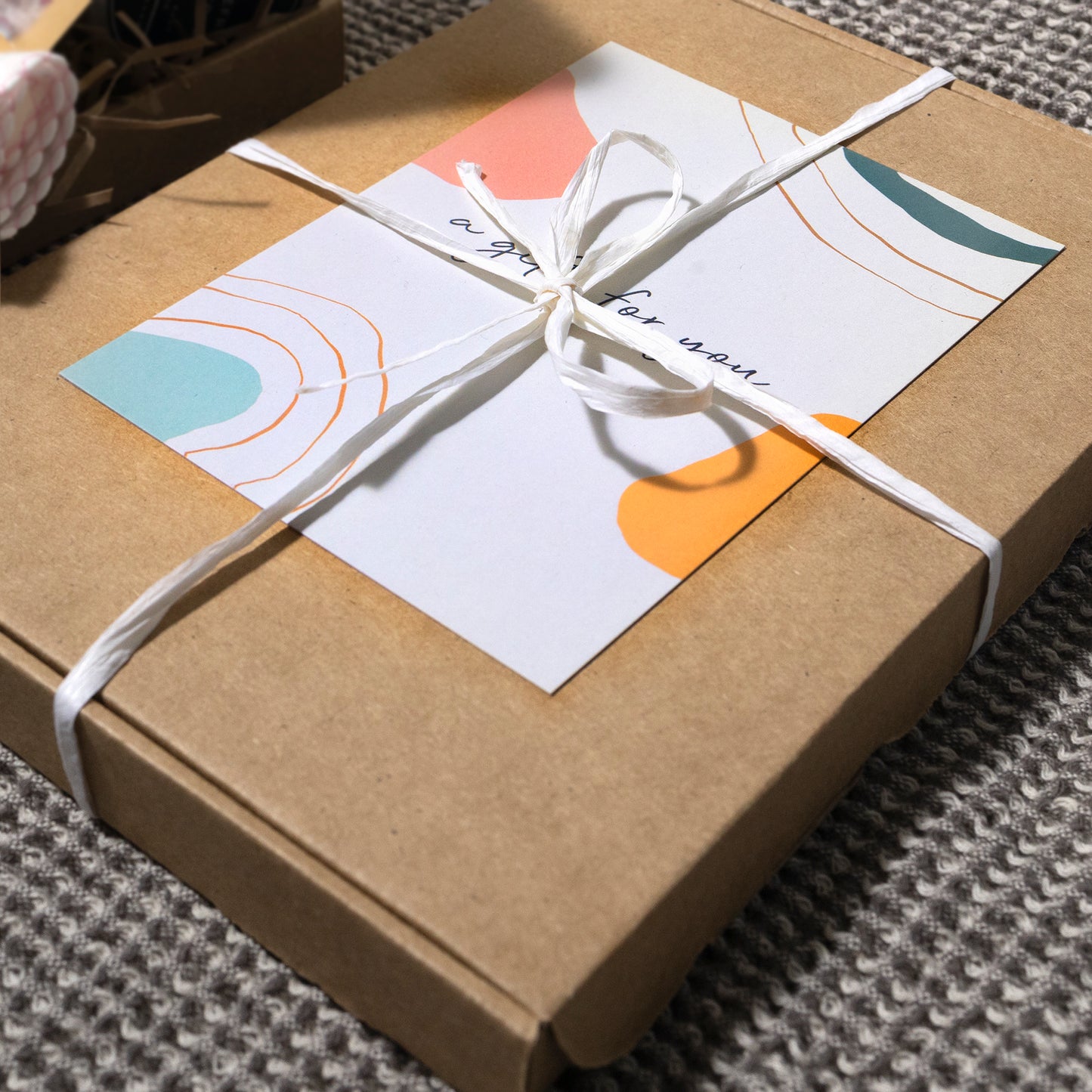 A cardboard gift box tied with a white string bow, topped with a card featuring abstract pastel art and handwritten text "A gift for you". The box is placed on a herringbone patterned fabric.