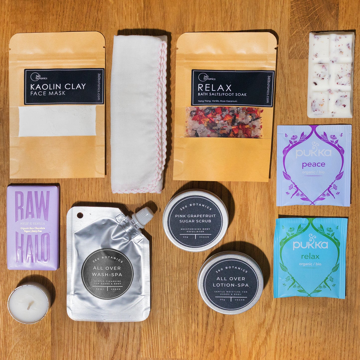 Relax bath salts & Kaolin clay face mask in kraft pouches, white muslin cloth, 2 tea bags, candle melts, body lotion and sugar scrub in silver round tins with black labels, t-light candle, all-over wash in silver packaging, a chocolate bar on wood