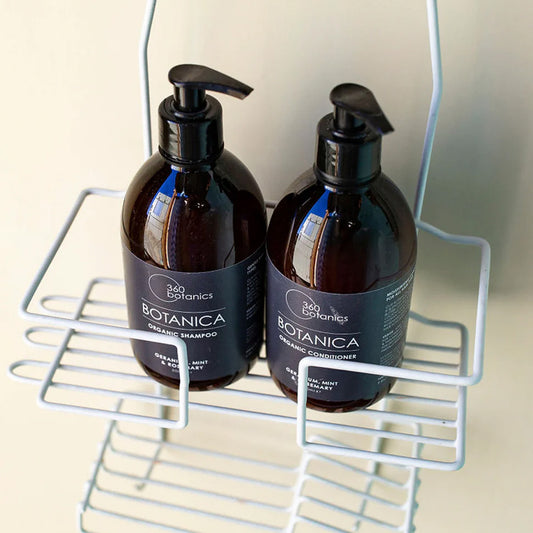 Two bottles of 360 Botanics Botanica Organic Conditioner neatly placed in a white metal shower caddy. The sleek, minimalist black packaging is highlighted by soft lighting, showcasing the Geranium, Mint, and Rosemary formulation for natural hair care