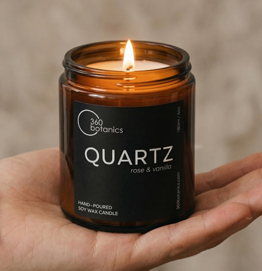 Hand holding a lit candle labeled 'Quartz' by 360 Botanics against a neutral background