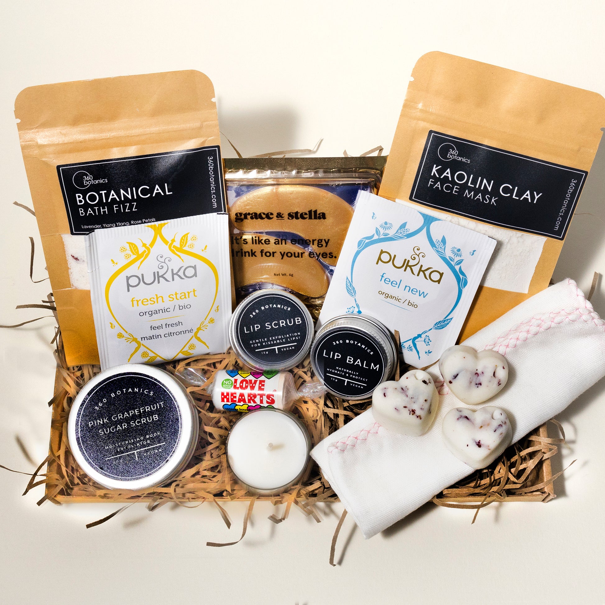 A gift basket containing a variety of self-care products including "Botanical Bath Fizz," "Kaolin Clay Face Mask," herbal teas from Pukka, "Grace & Stella" eye masks, lip scrub and balm, a pink grapefruit sugar scrub, a candle, a face cloth, and heart-shaped bath bombs, all nestled on a bed of shredded cardboard
