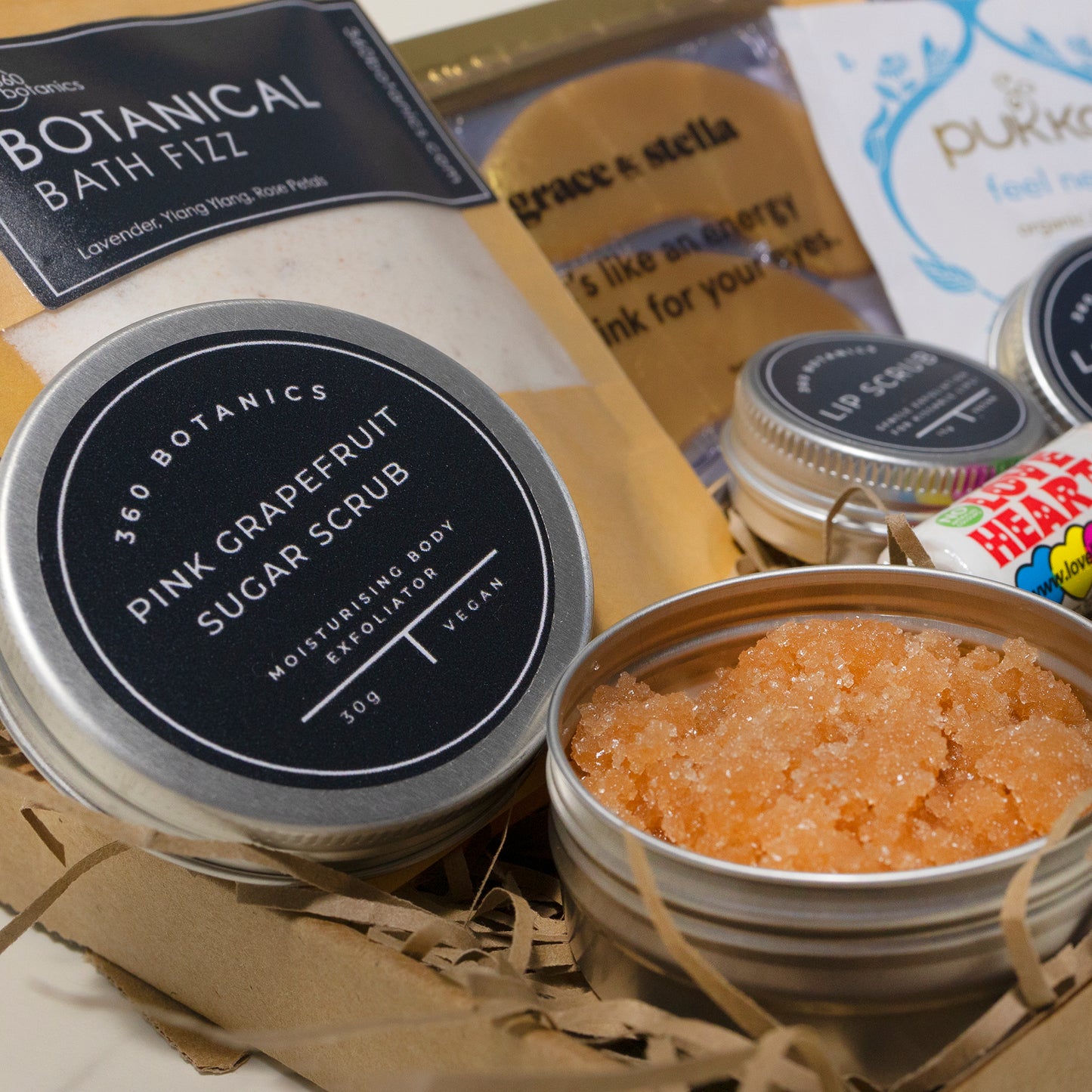  Close-up of "360 Botanics Pink Grapefruit Sugar Scrub" in an open metal tin, with granular texture visible, alongside other self-care products like "Botanical Bath Fizz" and "Grace & Stella" eye masks, partially visible in the background. The products are placed on a straw-filled surface.