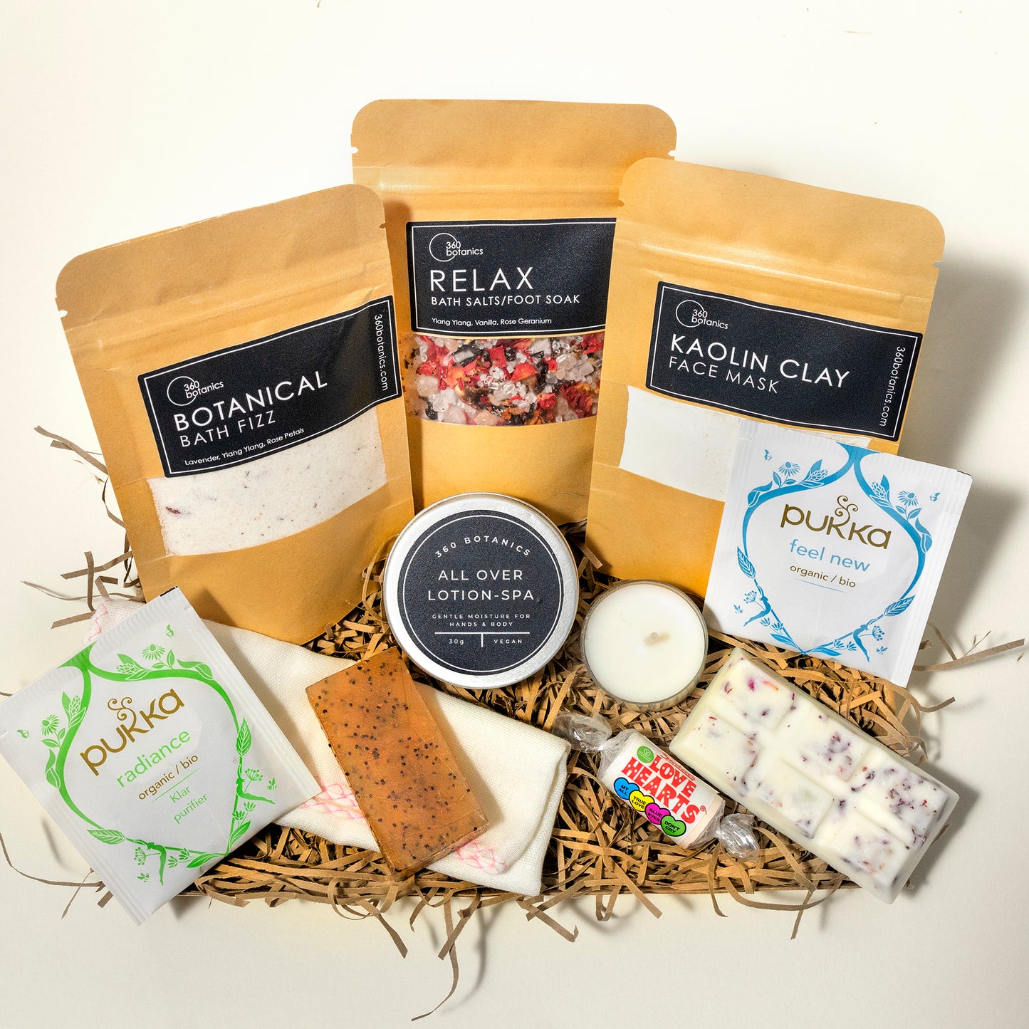 A variety of spa products arranged in a gift basket, including 360 Botanics Botanical Bath Fizz, Relax Bath Salts/Foot Soak, Kaolin Clay Face Mask, an All Over Lotion-SPA, a Pukka 'radiance' organic tea packet, a Pukka 'feel new' organic tea packet, a soap bar, and a candle, all nestled in natural wood straw.