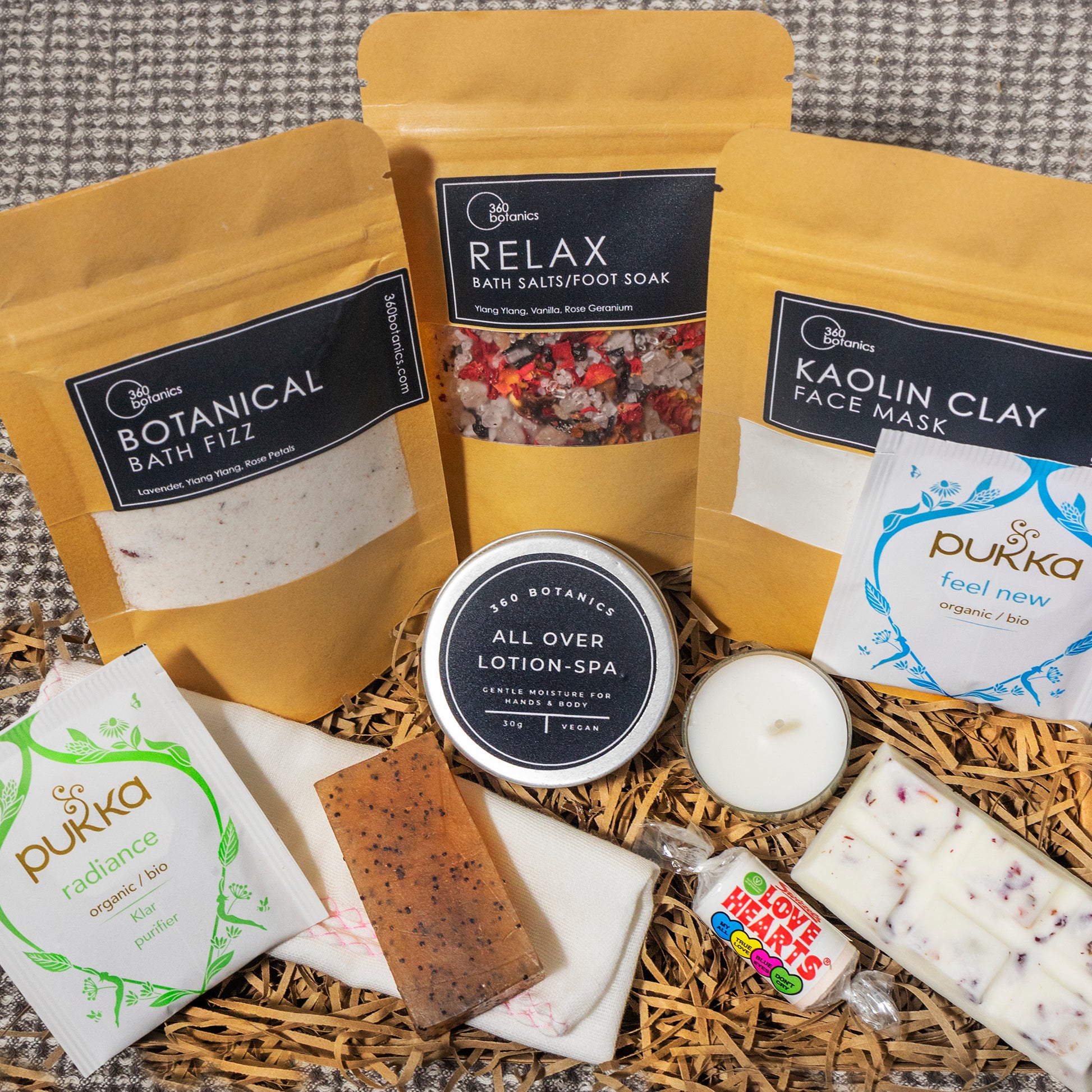 An assortment of bath and body products displayed on a herringbone patterned fabric. The collection includes 360 Botanics Botanical Bath Fizz, Relax Bath Salts/Foot Soak, Kaolin Clay Face Mask packets, an All Over Lotion-SPA in a tin, Pukka herbal tea packets in 'radiance' and 'feel new' varieties, a bar of exfoliating soap, a small white candle, and a Love Hearts candy roll, all presented on a bed of natural wood straw.