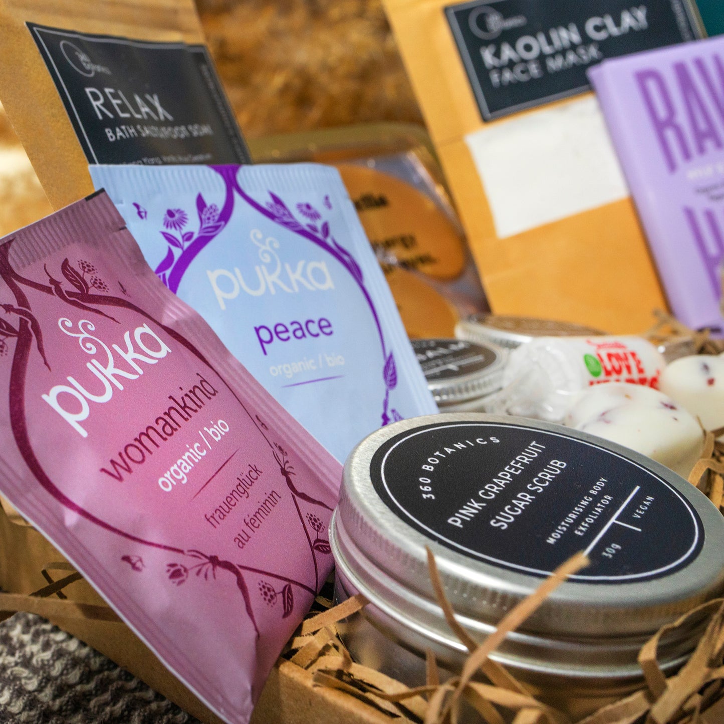 An angled view of a 360 Botanics wellness gift set showcasing a variety of products. In the foreground is a tin of Pink Grapefruit Sugar Scrub alongside sachets of Pukka organic teas in 'Womankind' and 'Peace' blends. Part of a 'RELAX Bath Salts/Foot Soak' pouch and 'KAOLIN CLAY Face Mask' are visible in the background, all artfully arranged in a rustic straw-filled box