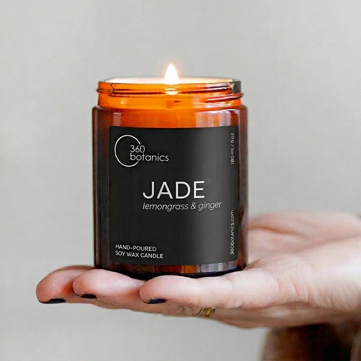 360 Botanics Lemongrass & Coriander Soy Wax Candle in an amber glass jar, gently held in a hand wearing a cozy knit sweater. The candle's flame glows softly, emphasising warmth and relaxation, ideal for creating a serene atmosphere