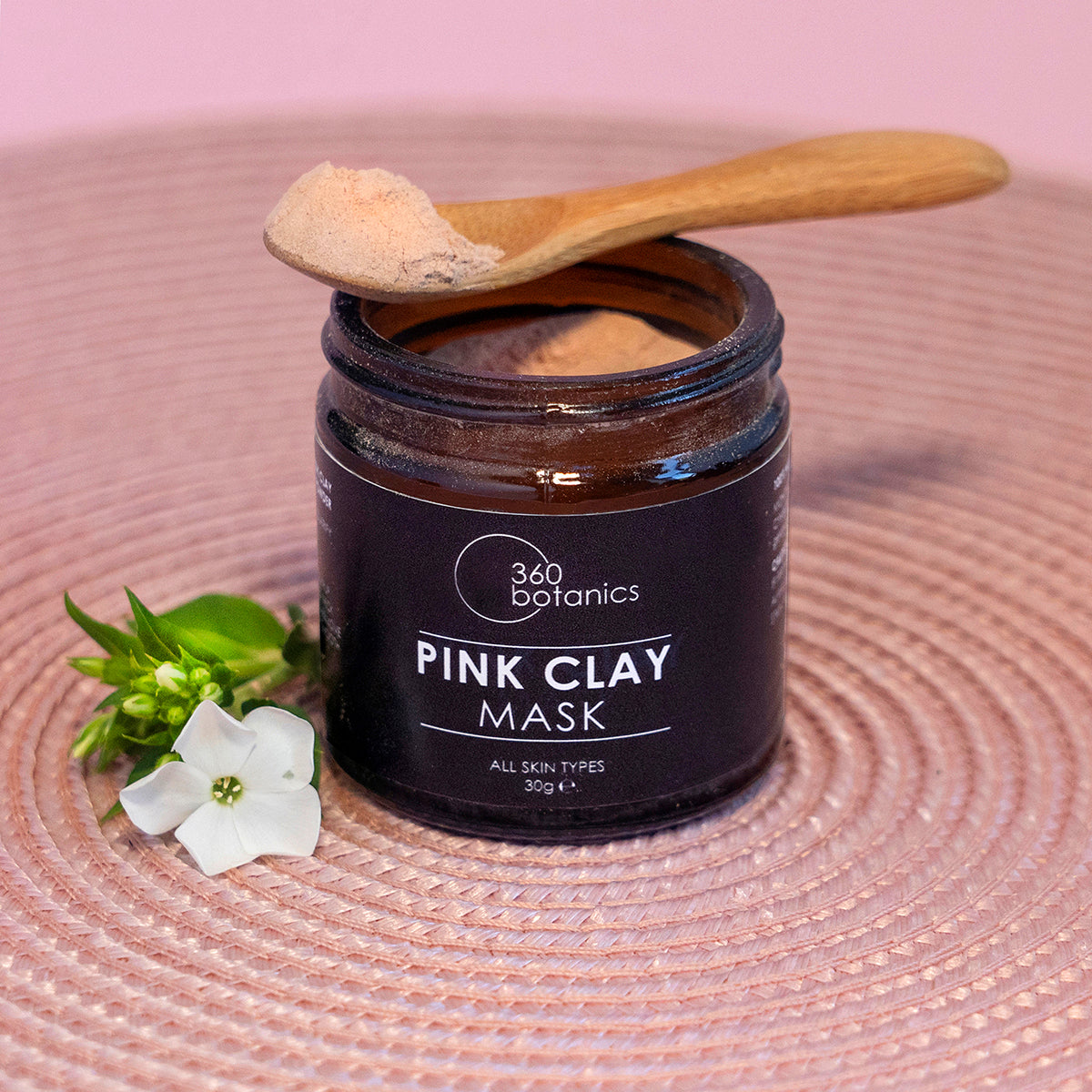 A jar of 360 Botanics Pink Clay Mask open on a woven mat, with a wooden spoon of product on top and a white flower accent, suggesting natural and organic skincare.