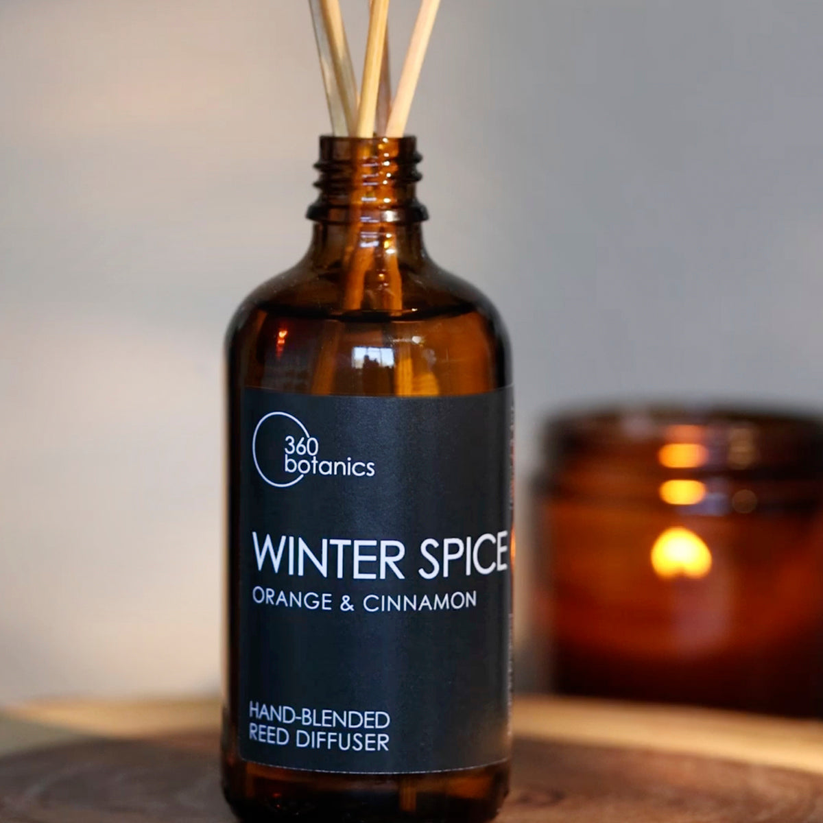 The 360 Botanics Winter Spice reed diffuser, featuring a blend of orange and cinnamon scents, is showcased in an amber glass bottle with reeds inserted. The warm glow of a candle in the blurred background adds a cosy atmosphere, highlighting the seasonal fragrance of the product