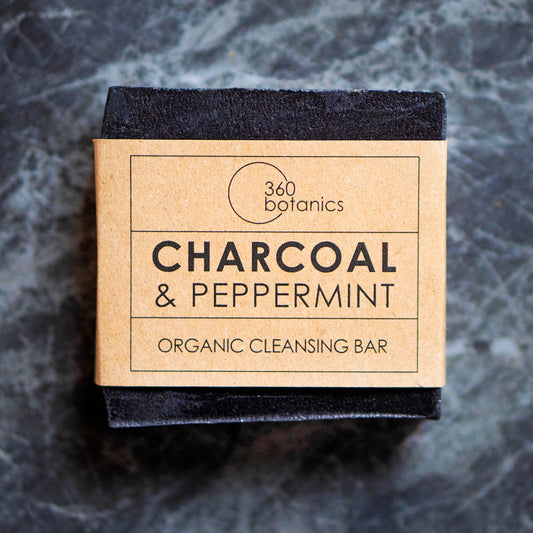 Close-up view of a 360 Botanics organic cleansing bar packaging, featuring 'CHARCOAL & PEPPERMINT' on a natural kraft paper label. The black bar of soap is partially visible, suggesting the product's natural charcoal ingredient, all set against a grey marbled background, evoking a clean and organic aesthetic