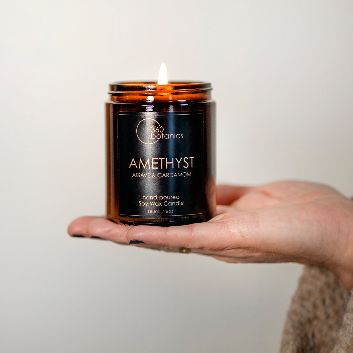 A hand holding a lit Amethyst Agave & Cardamom' scented soy wax candle by 360 Botanics, which is hand-poured into a brown glass jar.