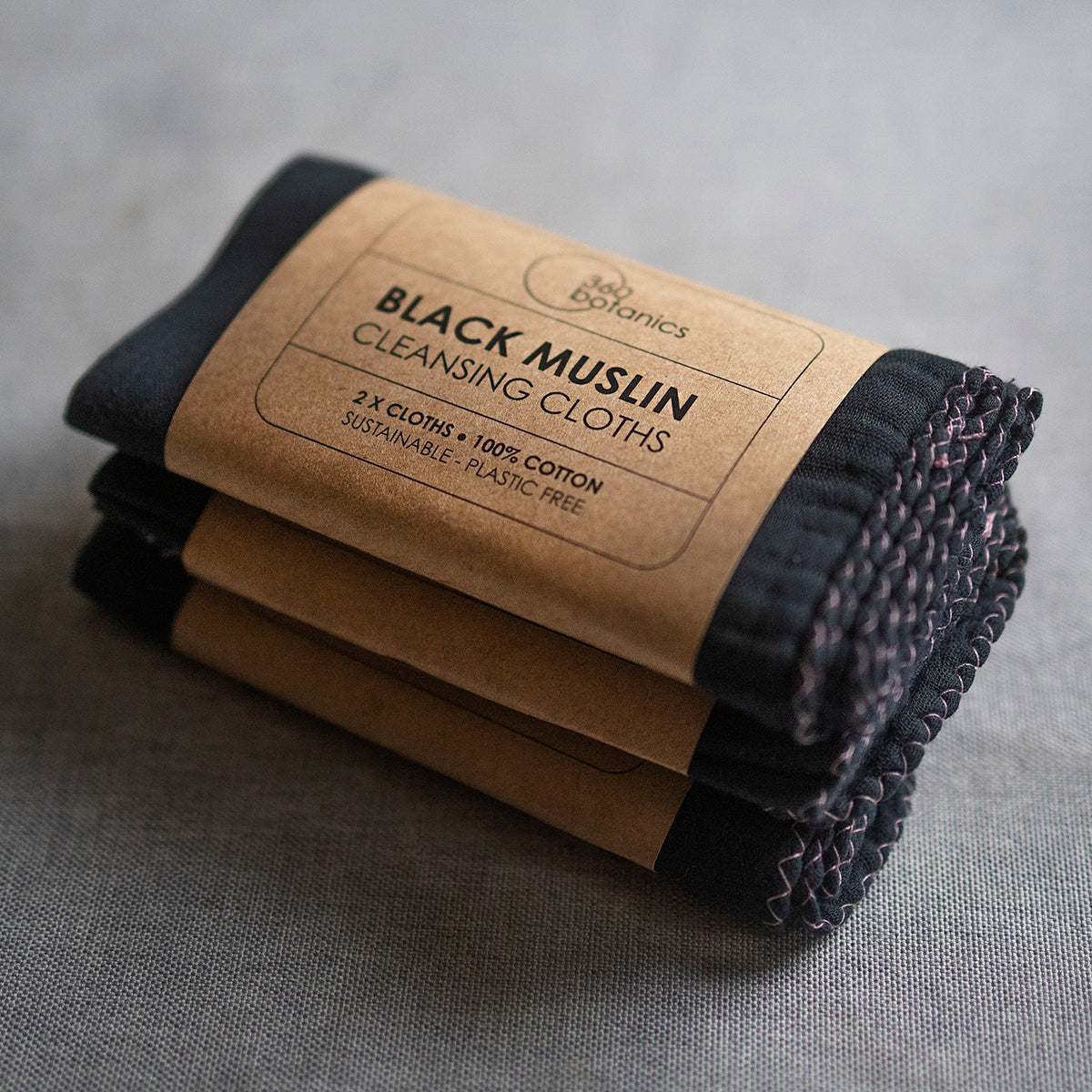  A stack of black muslin cleansing cloths, labeled as "2x Cloths, 100% Cotton, Sustainable, Plastic Free" by 360 Botanics, wrapped in a kraft paper band, set against a gray textured background.