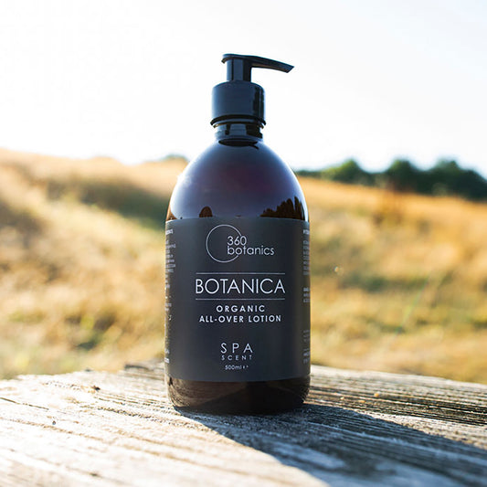 A 360 Botanics Botanica Organic All-Over Lotion bottle with a pump dispenser, in a 500ml size, resting on a wooden surface outdoors with a golden field in the background.