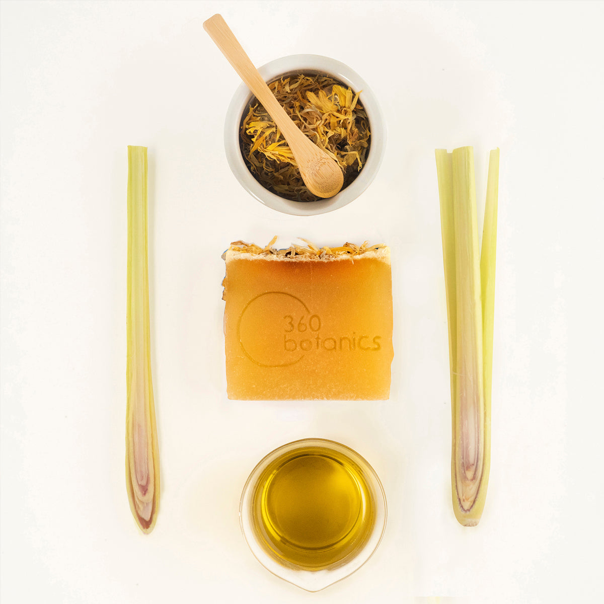 A flat lay composition featuring a 360 Botanics branded soap bar, a bowl of dried herbs with a wooden spoon, a small dish of oil, and two lemongrass stalks, all arranged on a white surface.