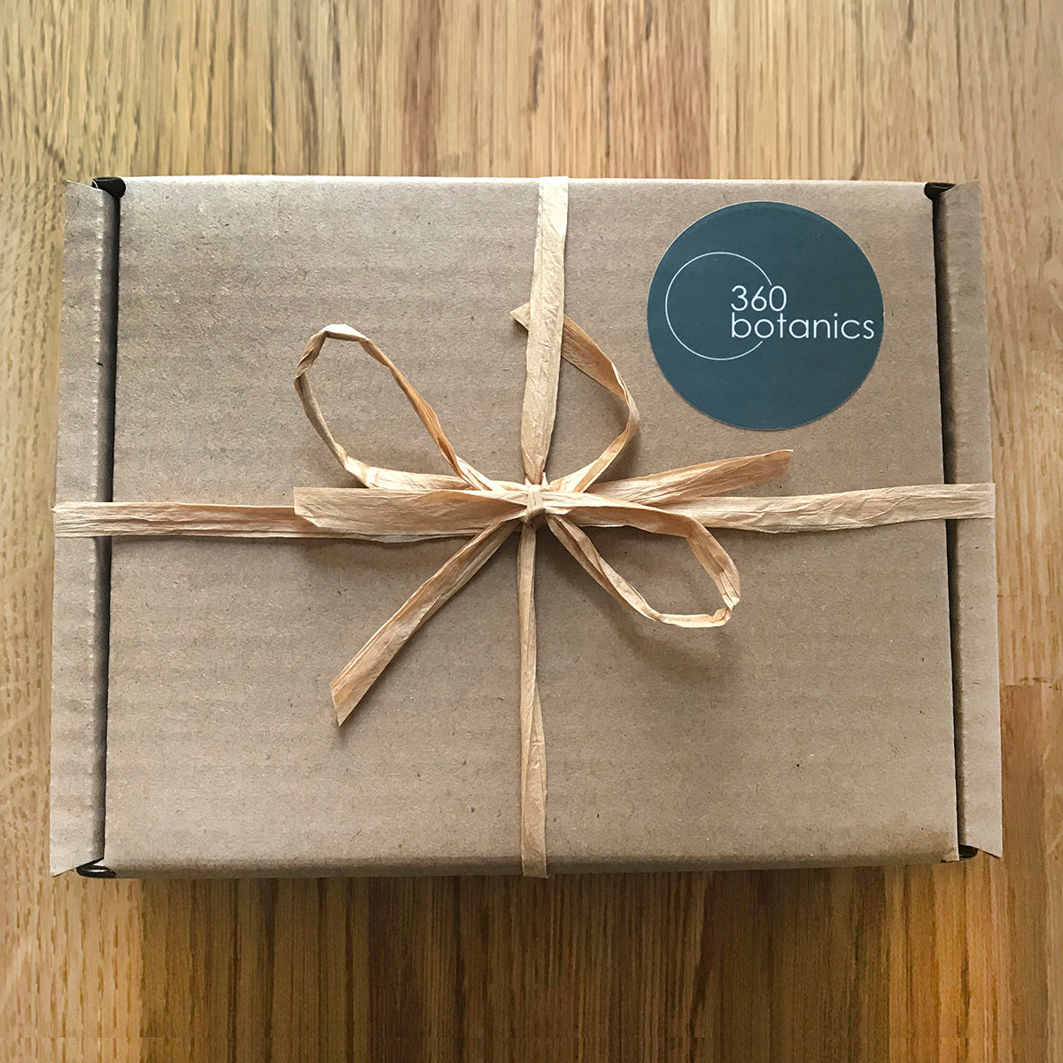 A closed cardboard gift box tied with a natural raffia ribbon and adorned with a circular "360 Botanics" sticker, presented on a wooden surface.
