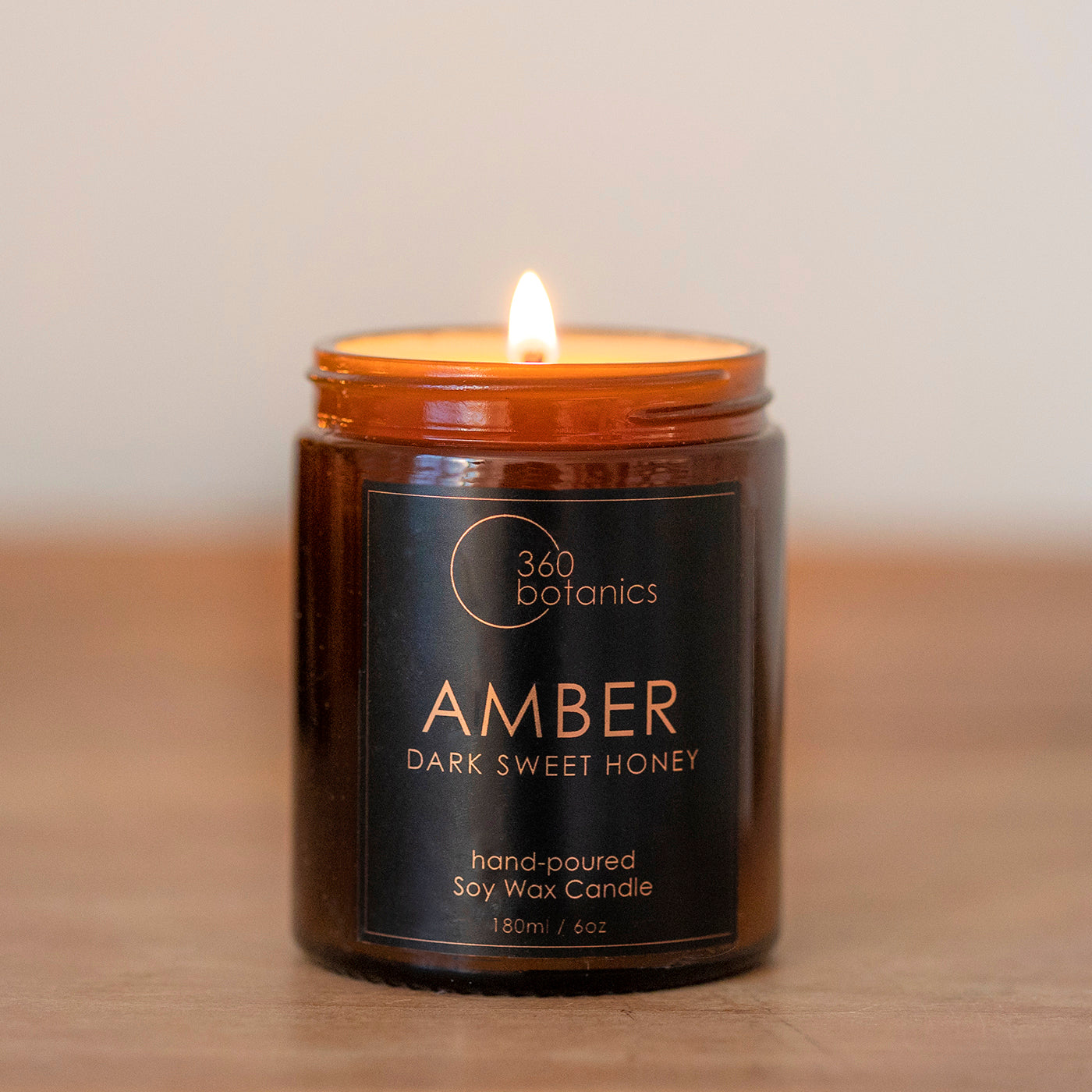 A lit 360 Botanics Amber hand-poured soy wax candle in a dark glass jar, labeled 'Dark Sweet Honey' with a visible flame, on a warm background.