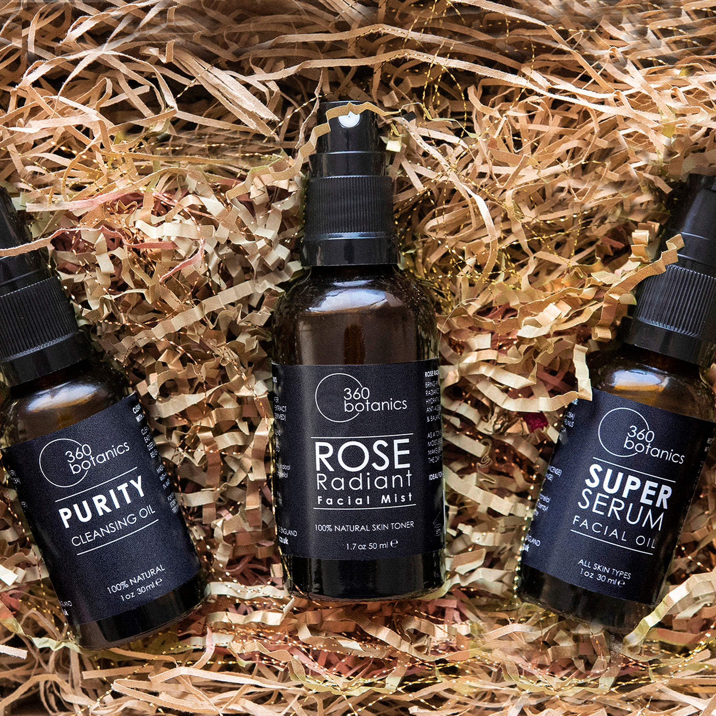 Three 360 Botanics skincare products nestled in shredded paper: Purity Cleansing Oil, Rose Radiant Facial Mist, and Super Serum Facial Oil, each labeled with their purpose and natural ingredients, in a 1 oz or 30 ml size.