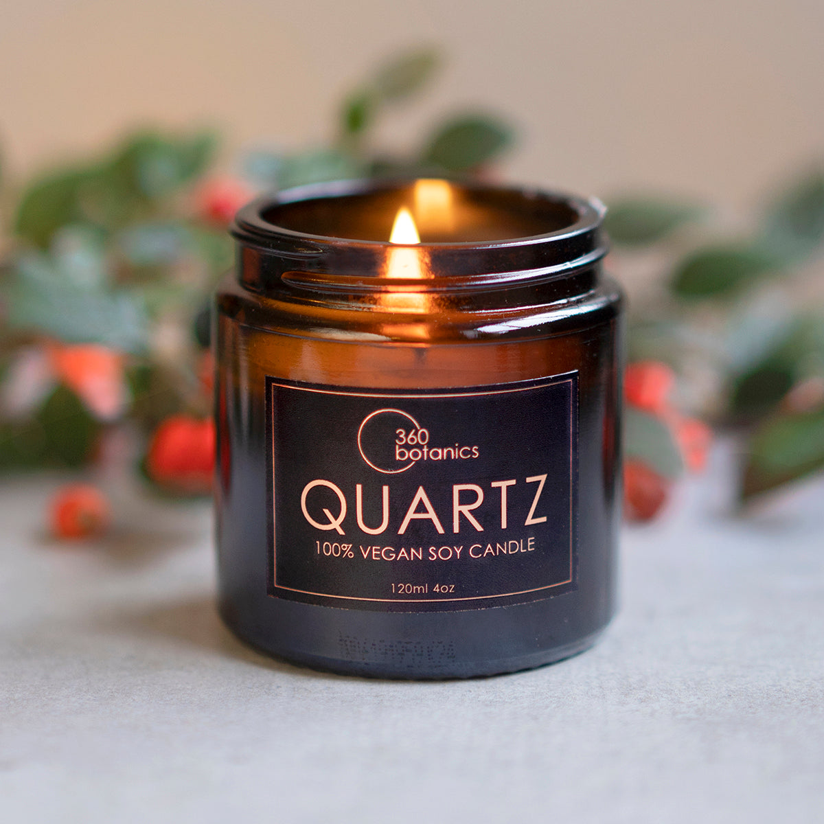 A lit 360 Botanics 'QUARTZ' vegan soy candle in a sophisticated black glass jar, with a gentle flame casting a warm glow. The 120ml jar is set against a soft focus background featuring subtle green foliage and hints of red berries, creating a serene and inviting atmosphere